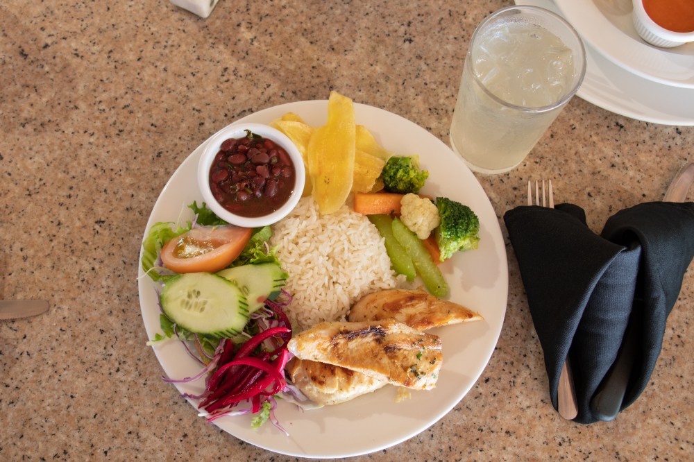 Plate with grilled chicken, rice, beans, salad, vegetables, chips, and iced drink on table.