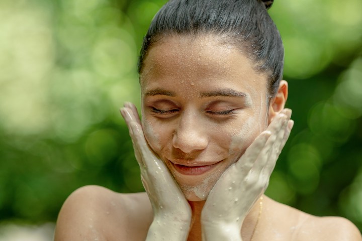 Person with clay mask on face, smiling with closed eyes, hands on cheeks, green background.