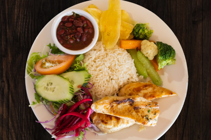Plate with grilled chicken, rice, beans, salad, plantain, and vegetables on a dark wooden table.