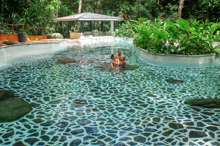 Two people relaxing in a patterned pool surrounded by lush greenery.