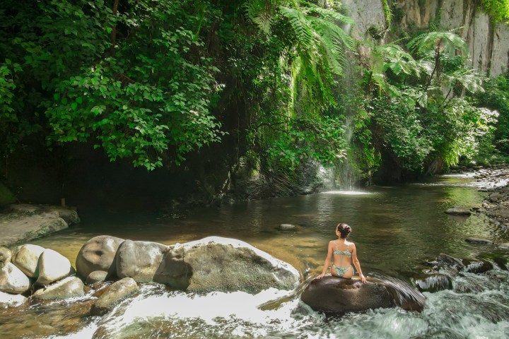 Person in swimwear sitting on rock in a waterfall pool surrounded by lush greenery.