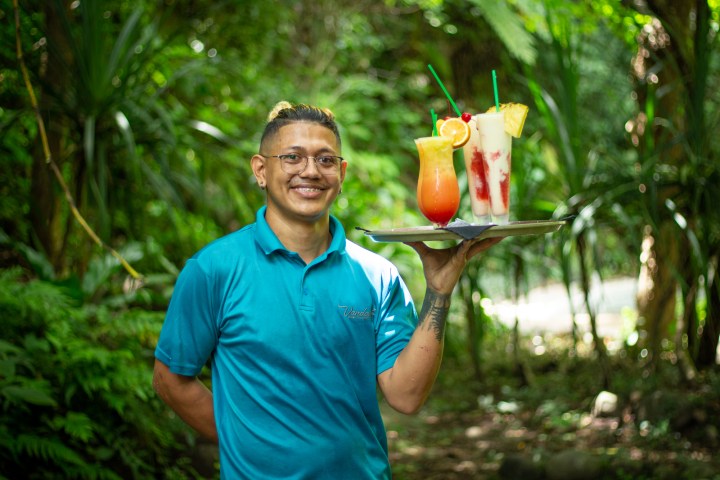 Smiling person in blue shirt holding a tray with three colorful drinks in a lush, green outdoor setting.