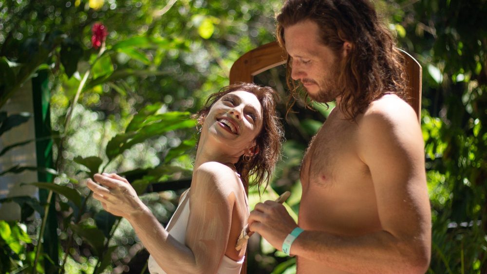 A woman smiles while applying lotion, standing with a man in a lush, tropical setting.