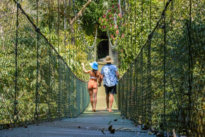 Two people walking on a rope bridge in a lush forest, wearing hats and summer clothing.