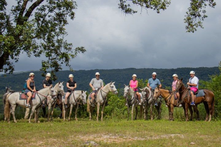Group of people on horses under cloudy sky with forested hills in background.