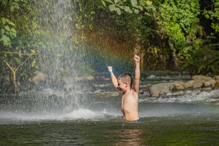 Child playing in a river under a waterfall with a rainbow, surrounded by greenery.