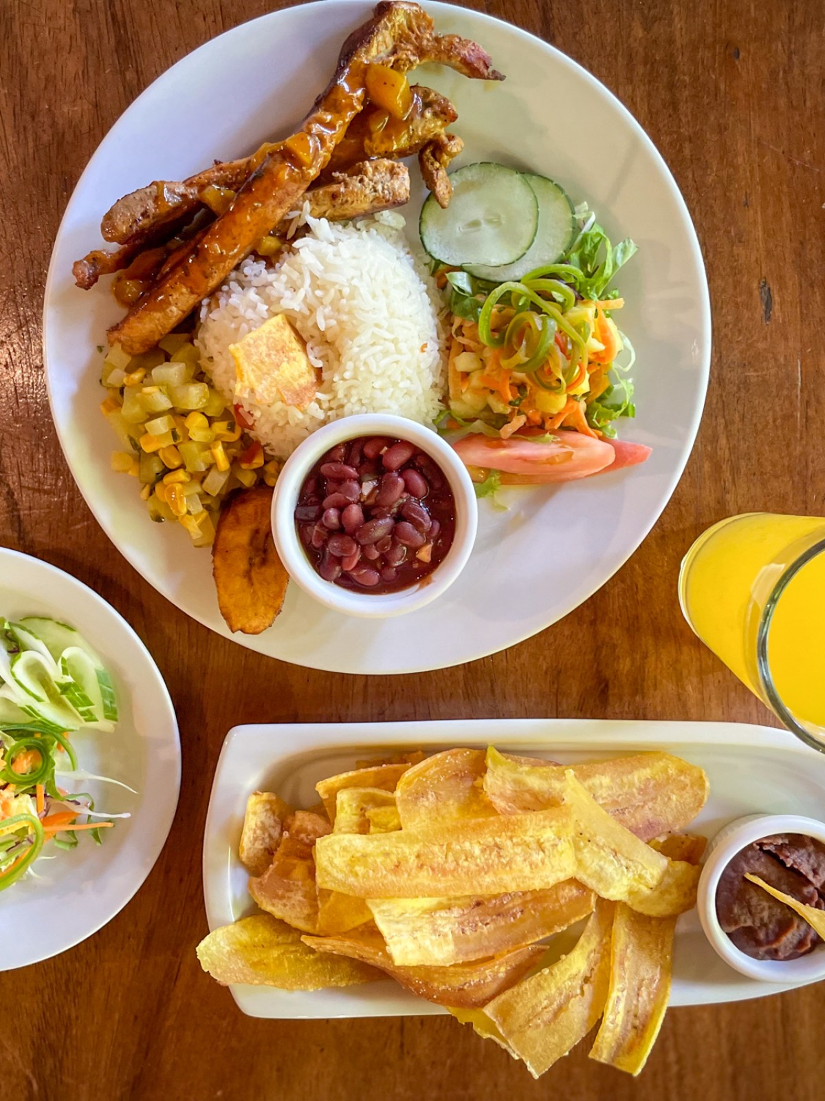 Assorted meal with rice, beans, meat, salad, plantains, and juice on a wooden table.