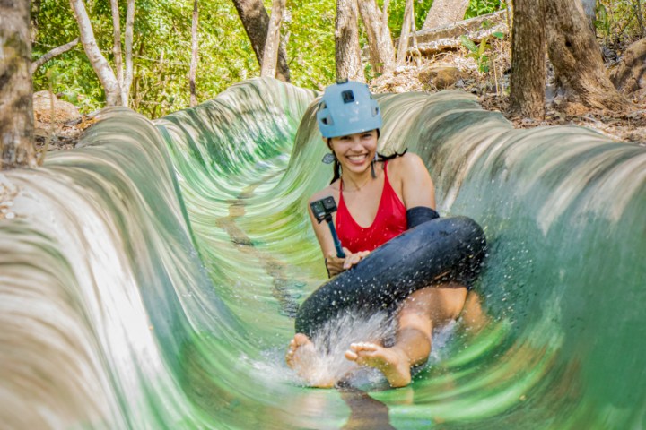 Person wearing helmet water tubing down a forest slide, smiling and holding a camera.