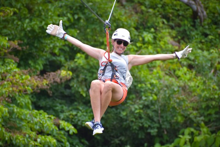 Person zip-lining in harness over green forest, arms outstretched, wearing helmet and sunglasses.