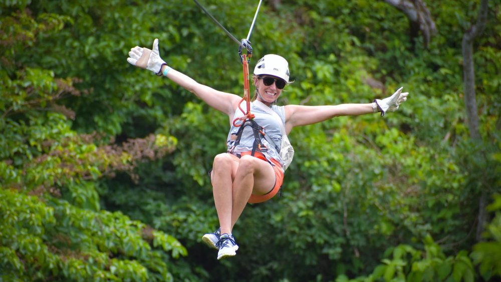 Person zip-lining in harness over green forest, arms outstretched, wearing helmet and sunglasses.