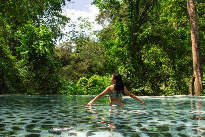 Person in a pool surrounded by lush green forest, under a partly cloudy sky.