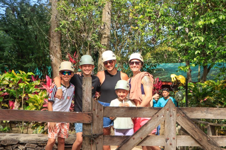 Group of five people wearing helmets, smiling outdoors near wooden gate and trees.