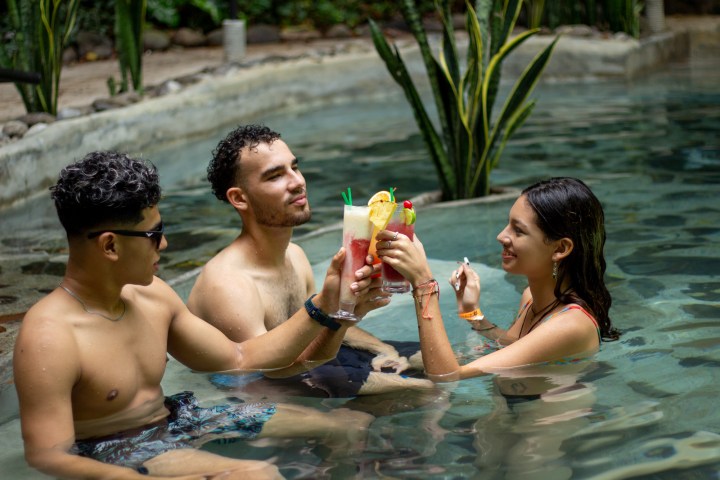 Three people in a pool enjoying colorful drinks and toasting.