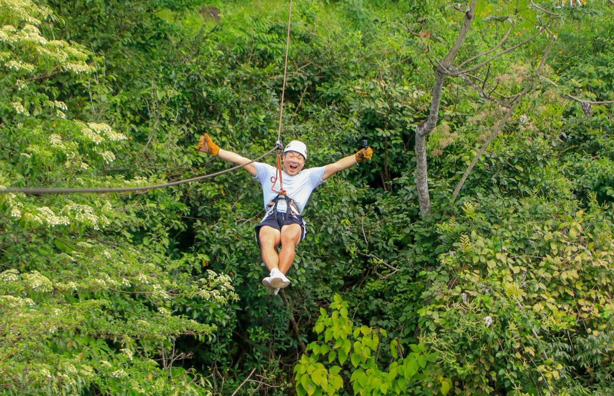 Person ziplining through lush green forest, arms outstretched, wearing helmet and gloves.