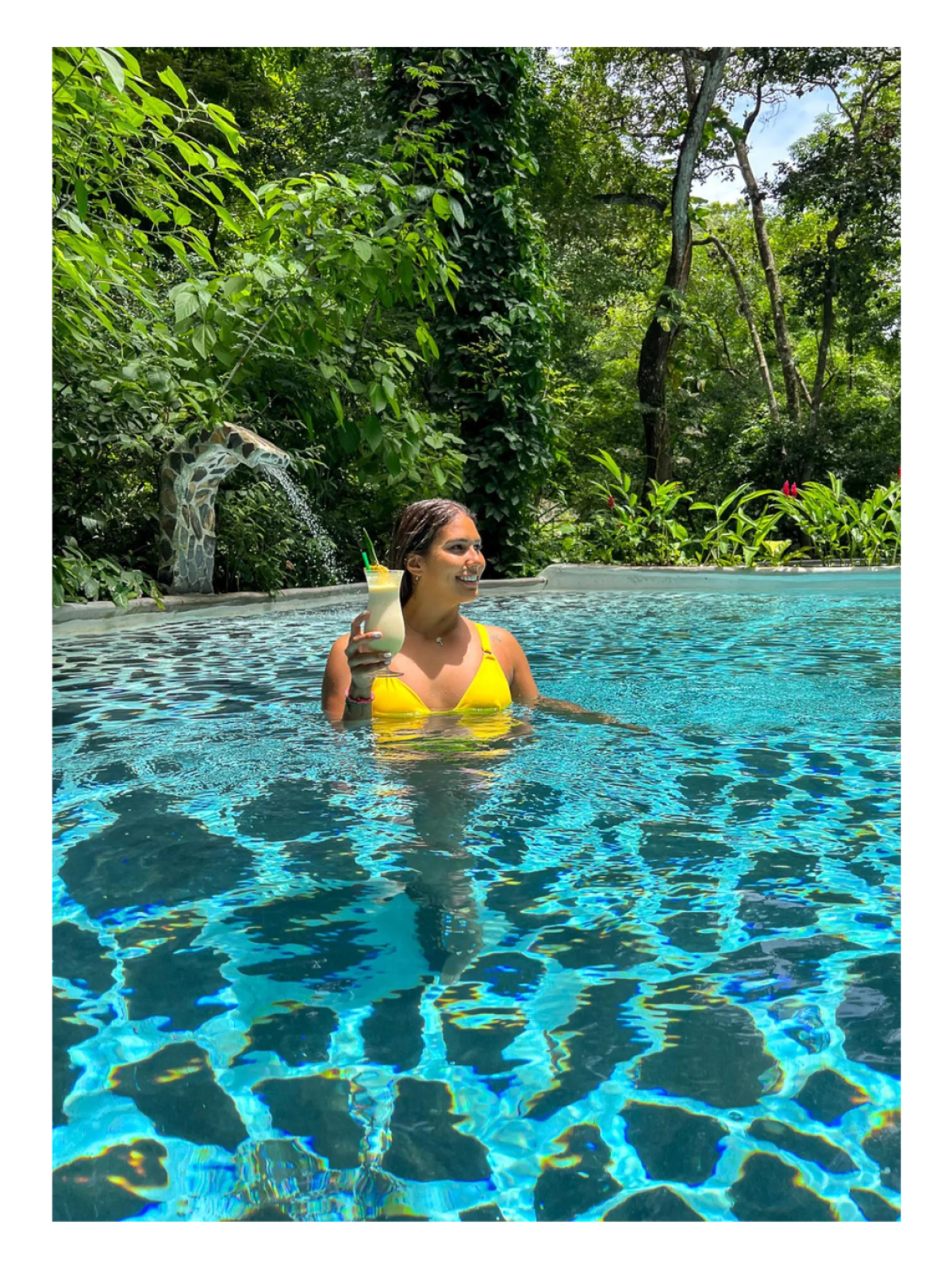 Person in yellow swimsuit holding a drink in a pool surrounded by lush greenery.