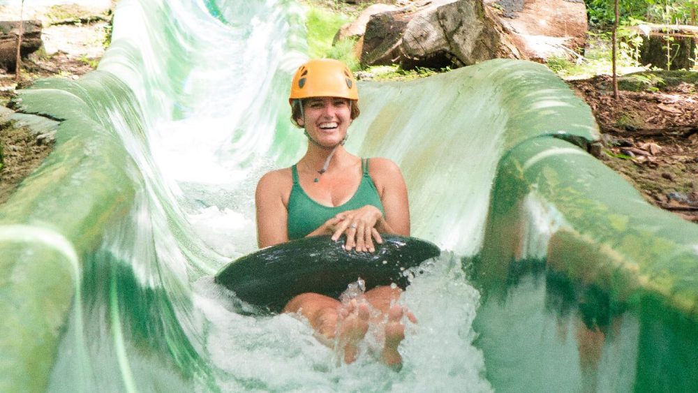 Person in green top tubing down a water slide in a forest setting, wearing a yellow helmet.