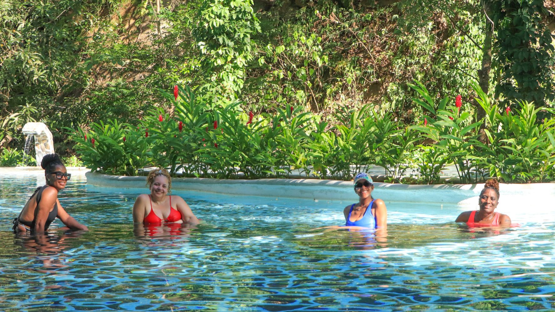 Four women in swimsuits enjoying a pool surrounded by lush greenery.