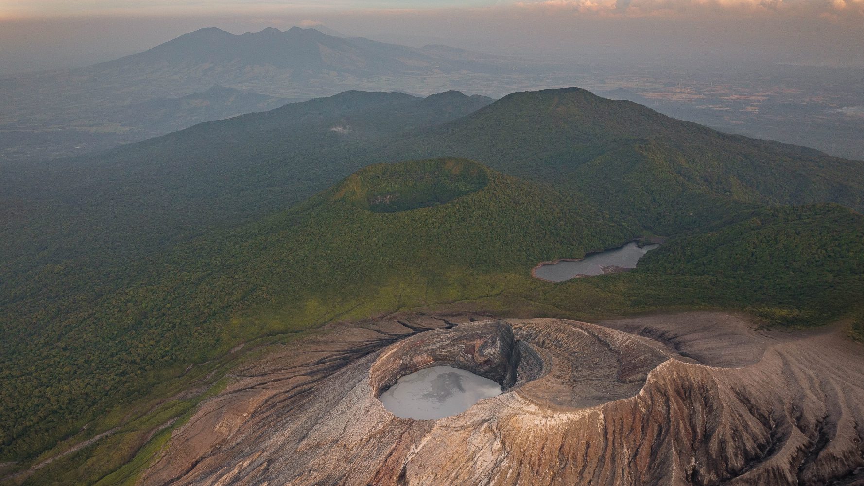 Aerial view of a volcanic crater with surrounding greenery and distant mountains under a cloudy sky.
