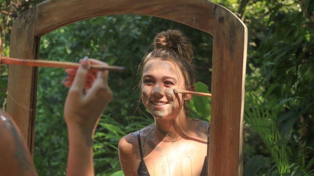 Person applying face paint in a mirror outside, surrounded by greenery.